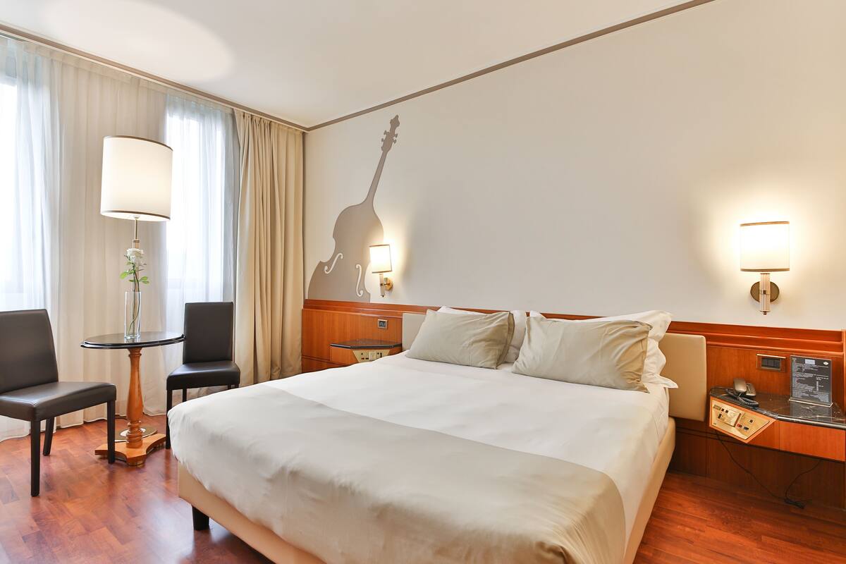 Une chambre d’hôtel avec un grand lit, deux chaises, une petite table avec une lampe, une décoration murale représentant une guitare et une table de chevet avec une lampe et un téléphone.
