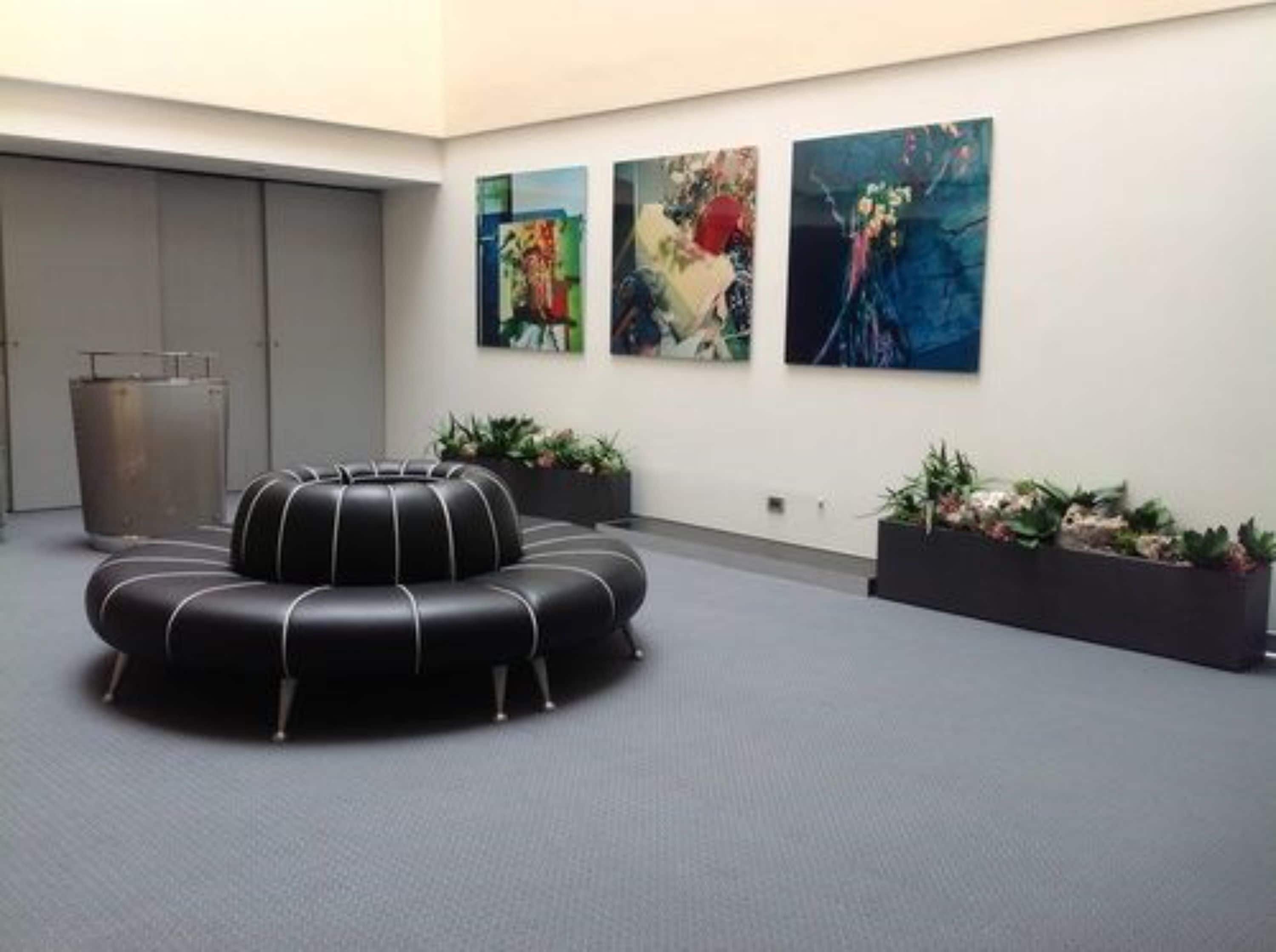 Un área de descanso moderna con un sofá circular de cuero negro, tres cuadros abstractos en la pared y dos macetas rectangulares con plantas.
