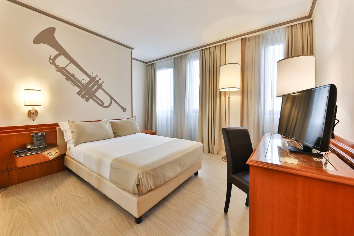 Une chambre d’hôtel avec un lit, un bureau avec une télévision, une lampe, une décoration murale représentant une trompette et une fenêtre avec des rideaux.