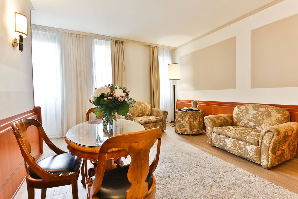 Une chambre d’hôtel comprenant une table à manger ronde, deux chaises en bois, un centre de table floral, un canapé, une table d’appoint et une lampe.