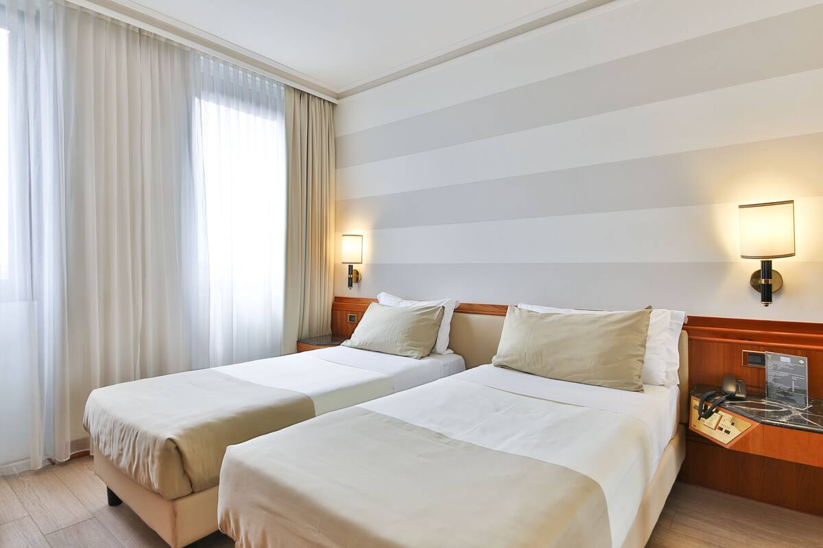 Une chambre d’hôtel avec deux lits, une tête de lit en bois, une table de chevet avec une lampe et une fenêtre avec des rideaux.