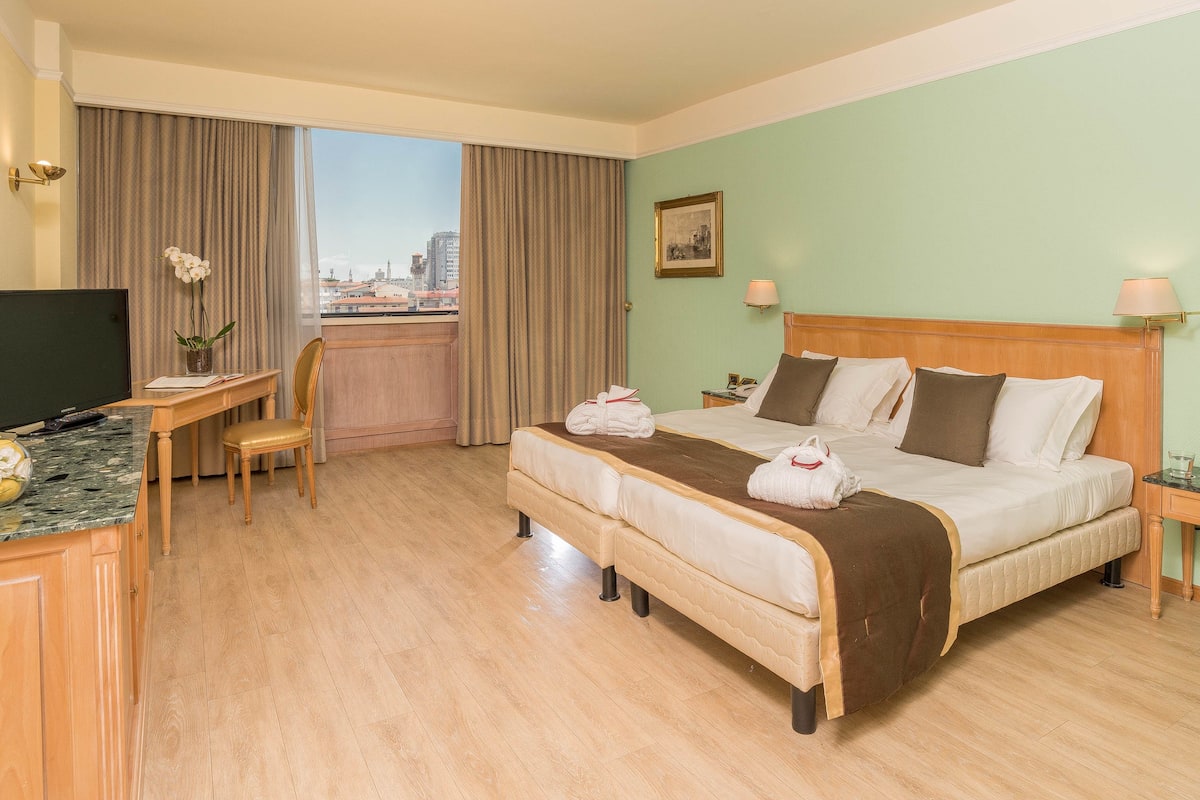 Een hotelkamer met een bed, een bureau met computer, een stoel, een televisie en een raam met gordijnen.