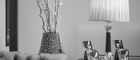 Una habitación con un sofá de cuero capitonado, un jarrón estampado con ramas, una mesa con una lámpara e imágenes enmarcadas, y un retrato en la pared.