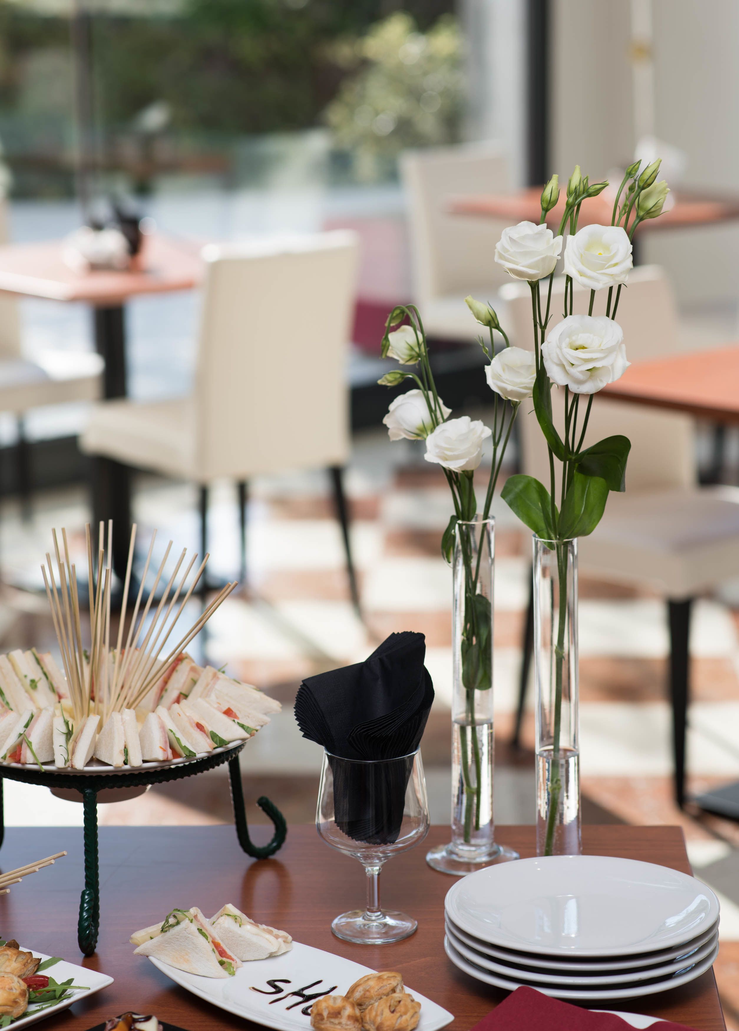 A dining table with a variety of food items, including sandwiches, pastries, and a plate with a napkin. There are also vases with white flowers and a glass with a black napkin.