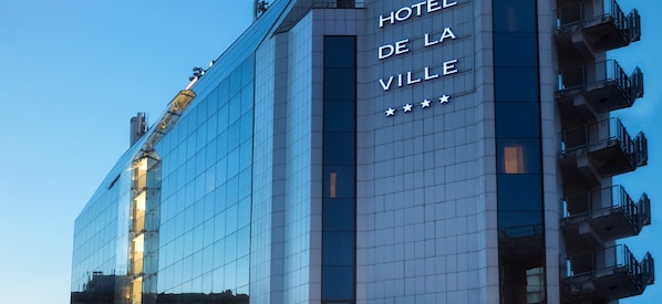 A modern hotel building with a reflective glass facade and a sign reading "HOTEL DE LA VILLE".