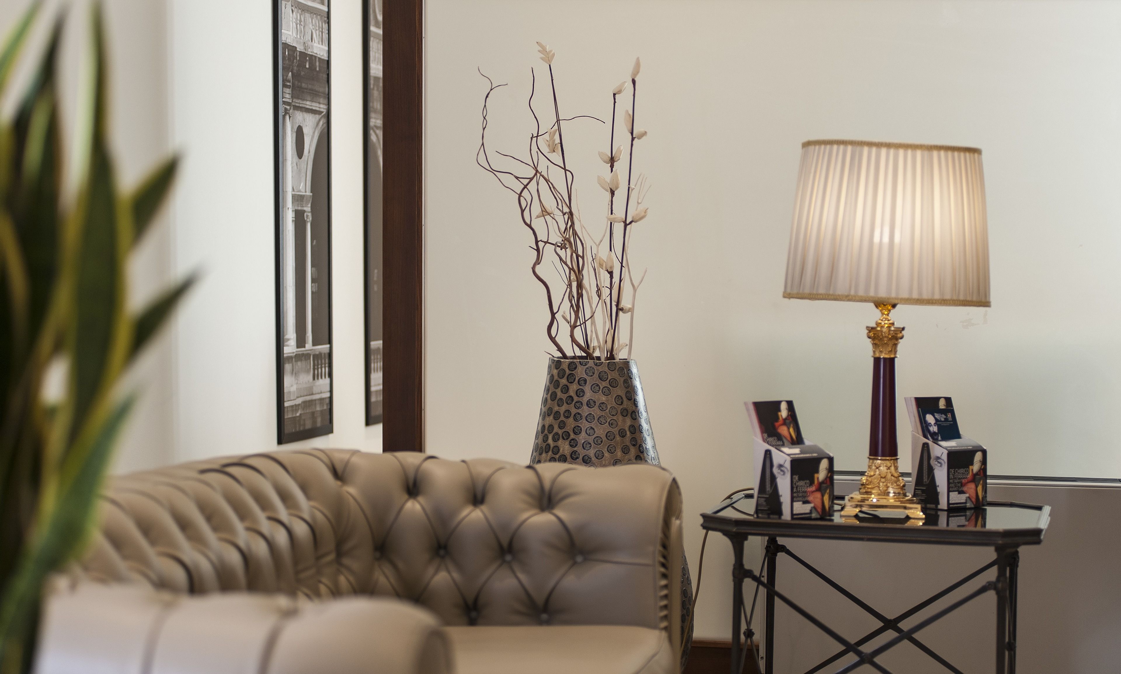 Een gestoffeerde leren bank, een vaas met takken, een tafel met een lamp en ingelijste foto's, en een ingelijste architectuurfoto aan de muur.