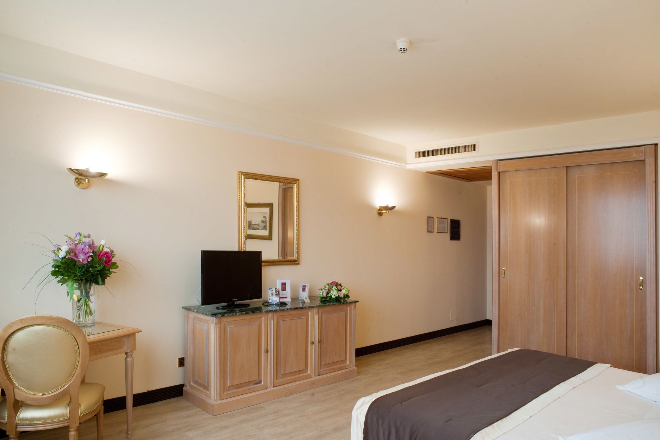 Una habitación de hotel con una cama, un armario de madera con un televisor de pantalla plana, un escritorio con un jarrón de flores y un guardarropa.