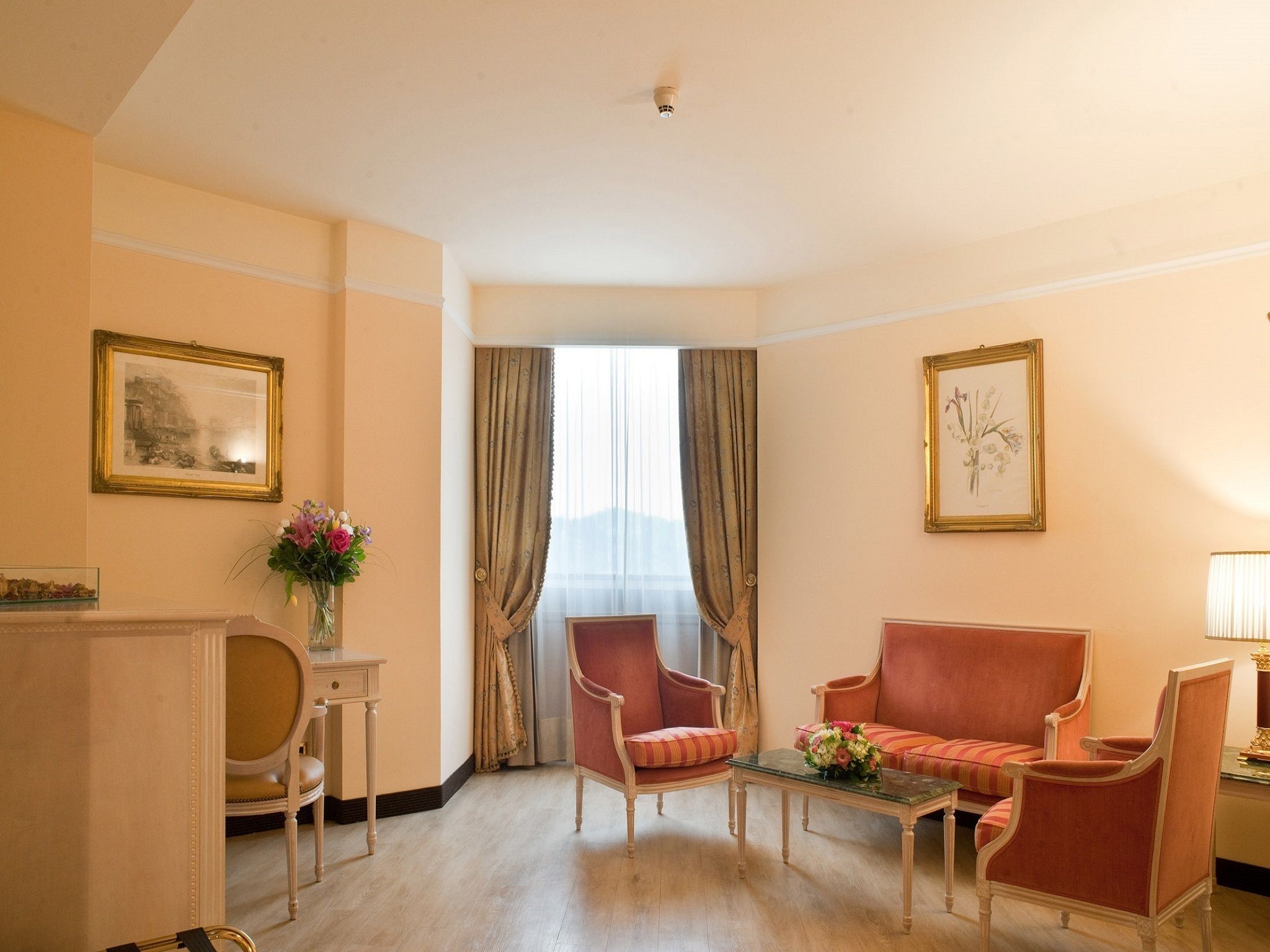 A hotel room with a sofa, two armchairs, a small table, a vase with flowers, and two framed pictures on the walls.