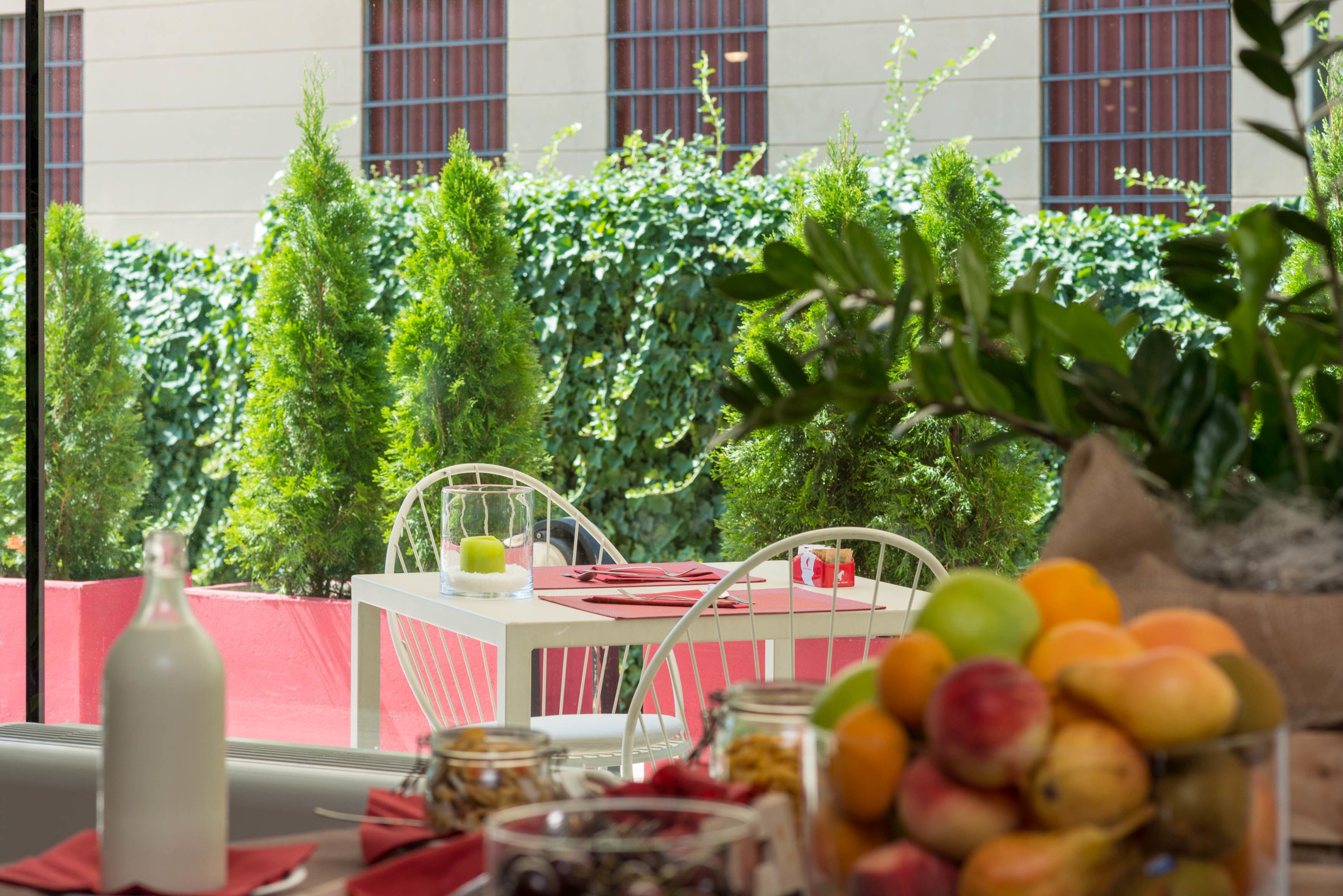 An outdoor dining area with a table set for two, surrounded by lush greenery and a building in the background.