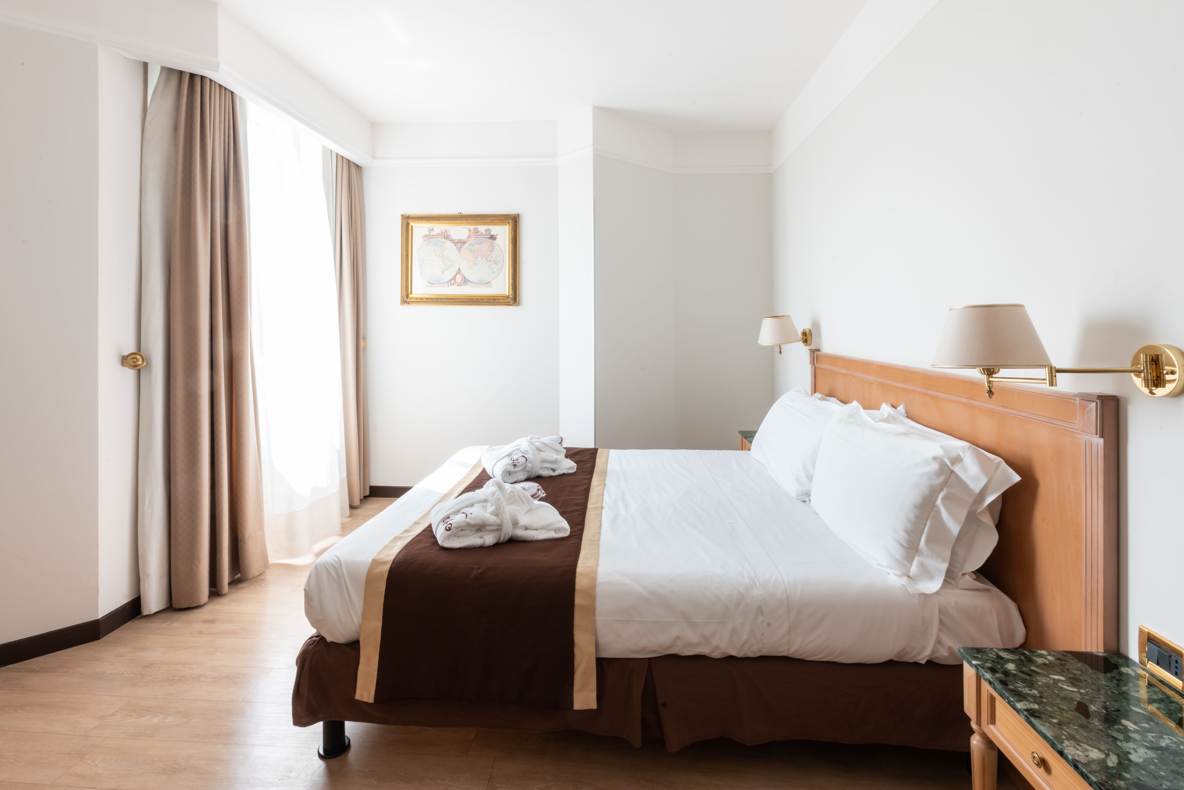 A hotel room with a neatly made bed, a wooden headboard, a nightstand, and a window with curtains.