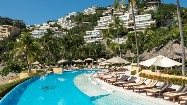 Piscina de resort com espreguiçadeiras, palmeiras e um conjunto de edifícios numa encosta.