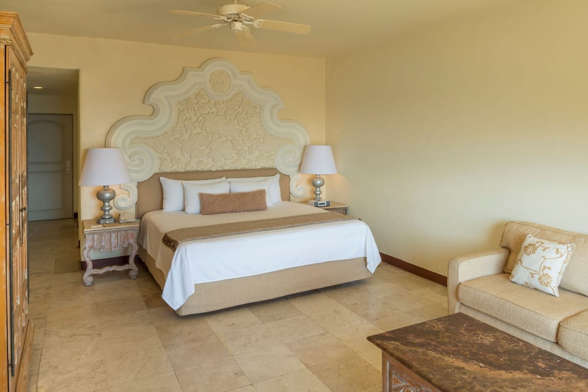 A hotel room with a large bed, two bedside tables with lamps, a ceiling fan, a sofa, and a wooden coffee table.