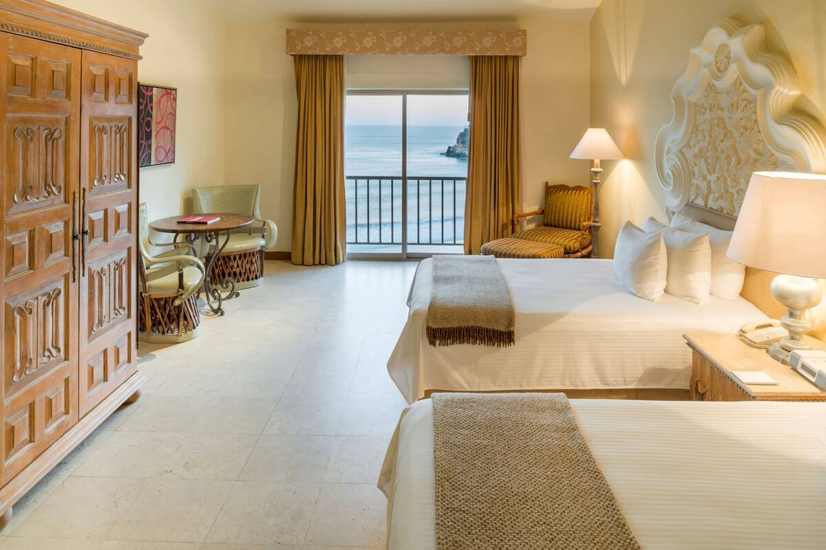 A hotel room with two beds, a balcony with ocean view, a wooden wardrobe, a small table with chairs, and a lamp.