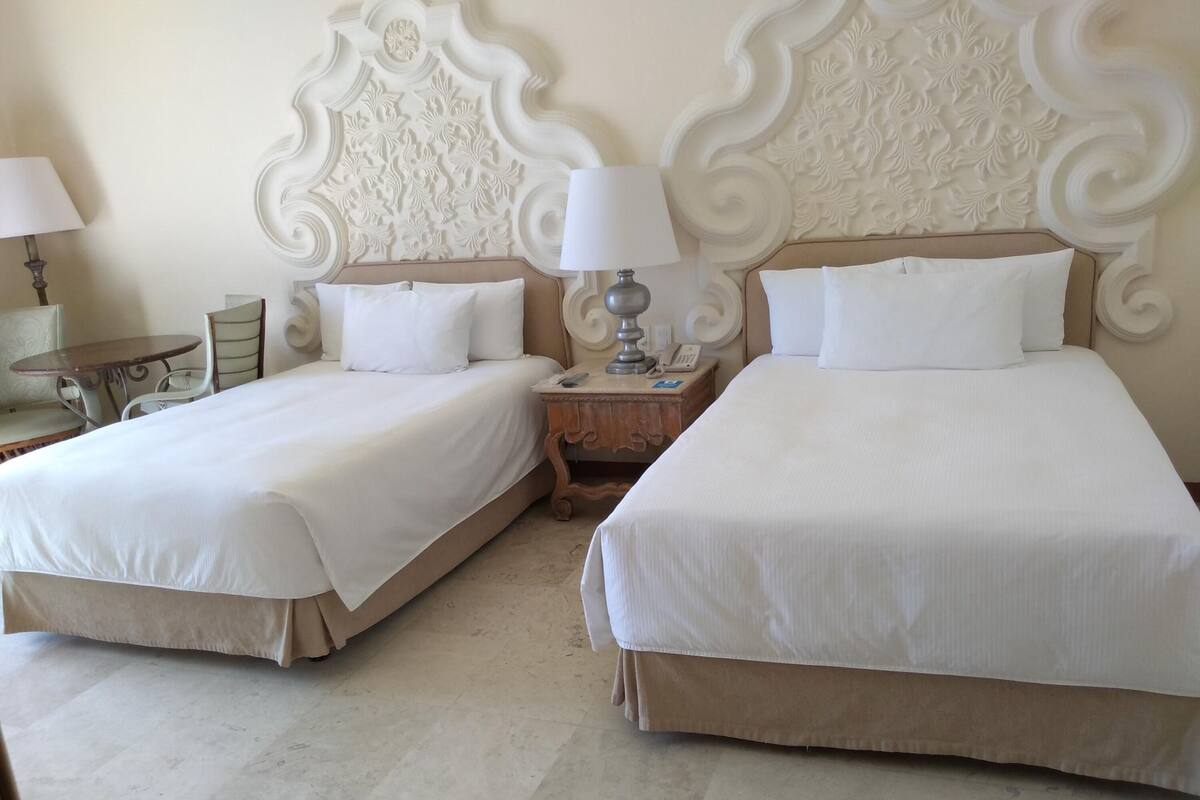 A hotel room with two beds, each with white bedding and a wooden headboard. There is a nightstand with a lamp and a phone between the beds.
