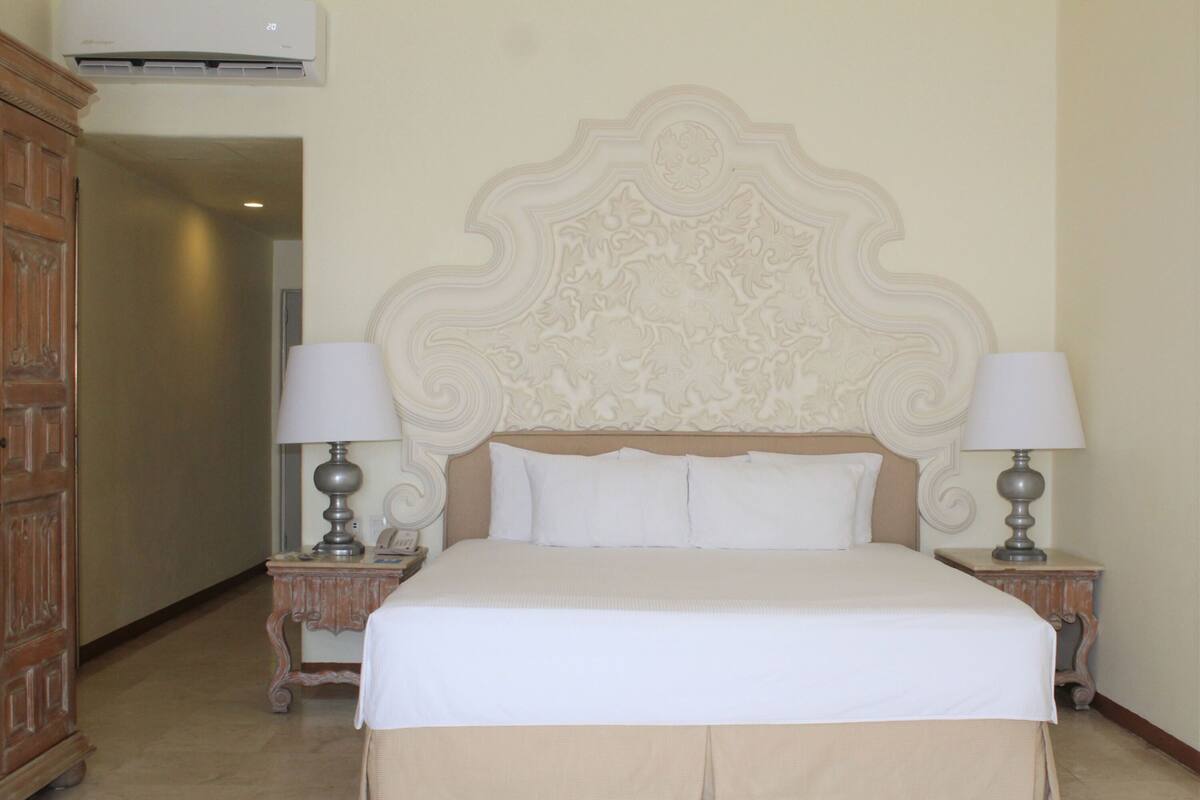 A hotel room with a large bed, two bedside tables with lamps, a wooden wardrobe, and a decorative headboard.