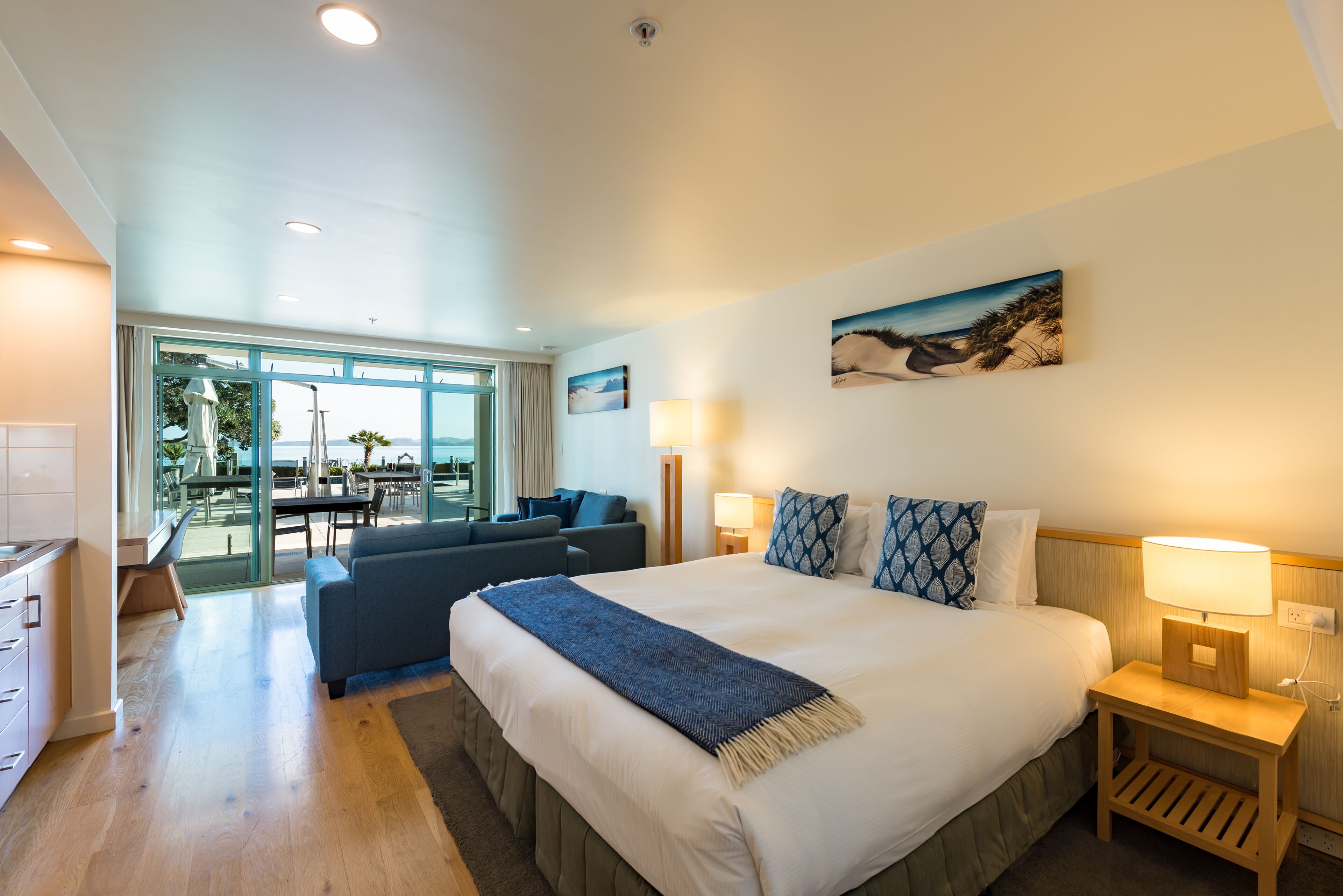 A modern hotel room with a large bed, a seating area with a sofa and chairs, and a view of the outdoors through large windows.