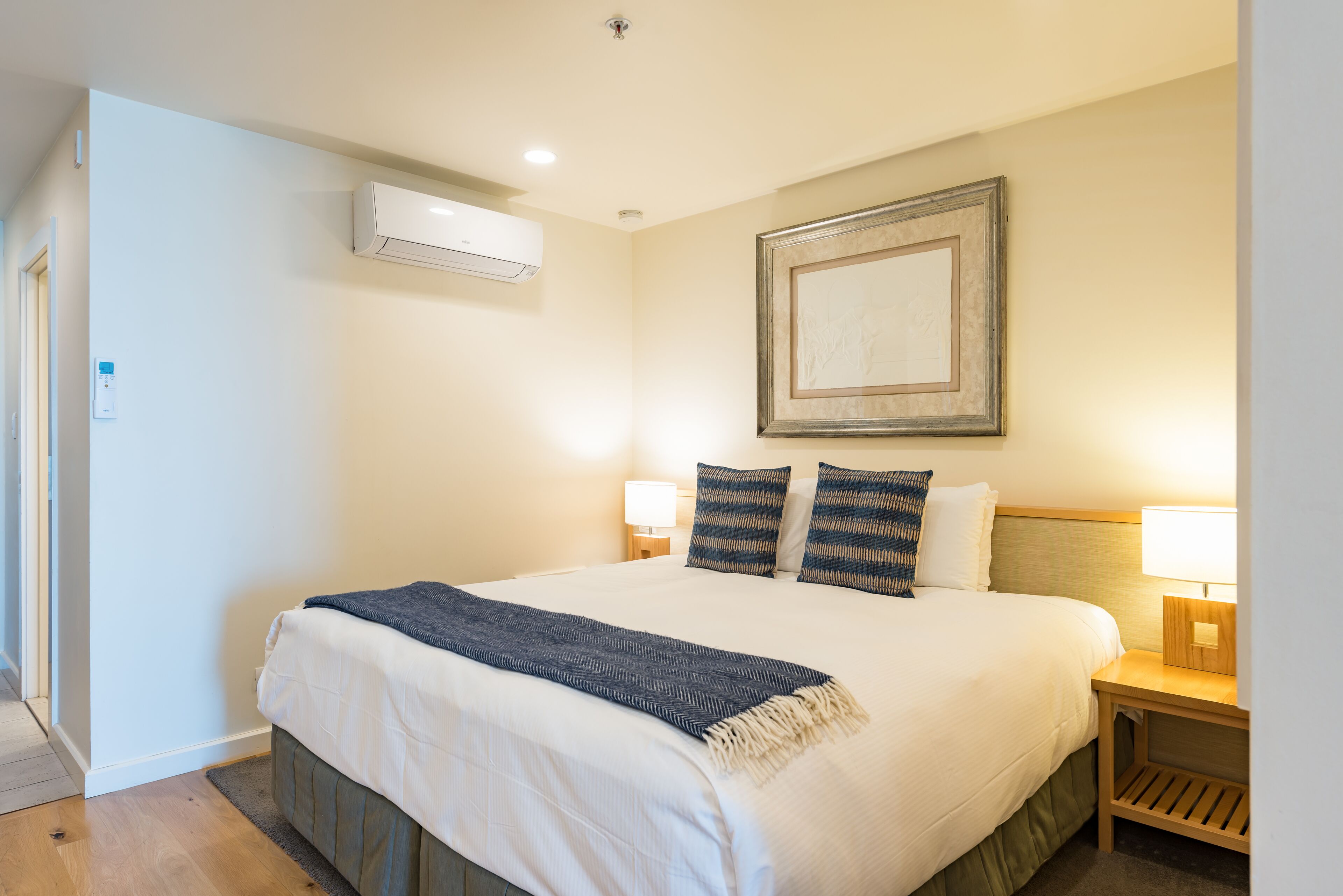 A neatly made bed with a dark throw blanket, two bedside lamps, a framed artwork above the bed, and an air conditioning unit on the wall.