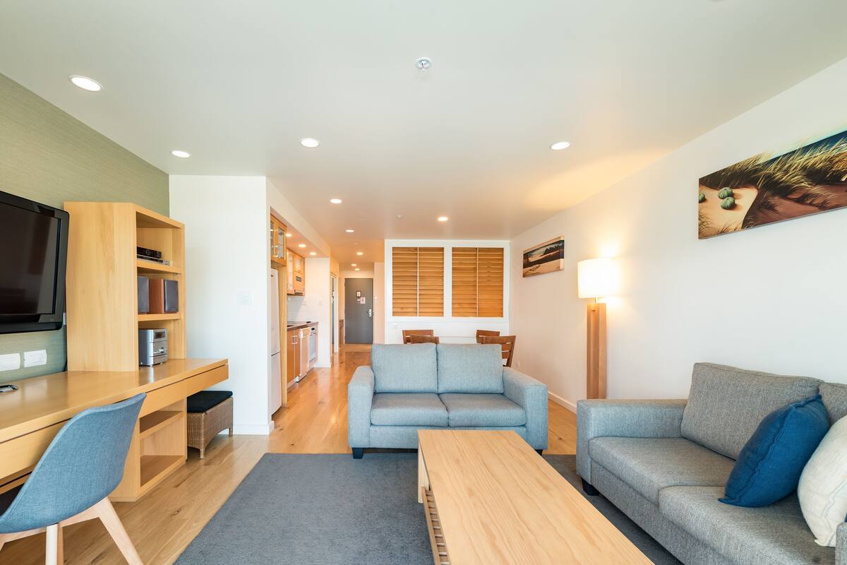 A modern living room with a sofa, a coffee table, and a TV. There is a kitchen area in the background.