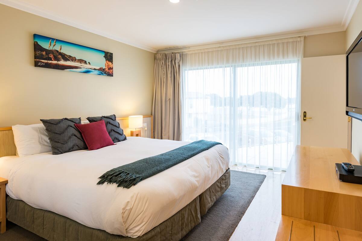 A hotel room with a large bed, a painting on the wall, a desk with a TV, and a window with curtains.
