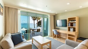 Een moderne woonkamer met een bank, een salontafel, een televisie en een bureau met een computer. Er is een glazen deur die leidt naar een balkon met uitzicht op zee en palmbomen.