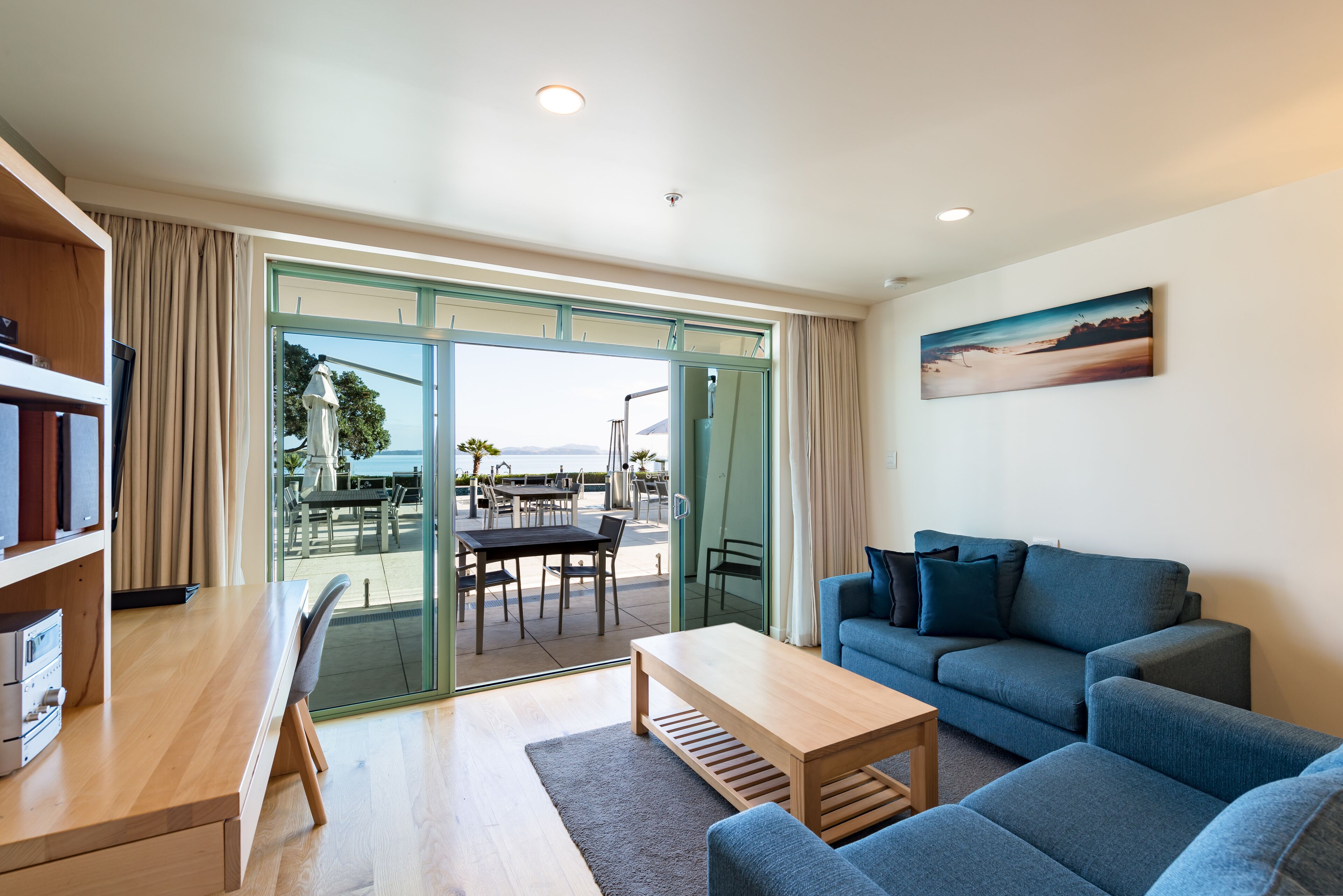 Una sala de estar moderna con un sofá, una mesa de centro y un escritorio con una silla. Una puerta de vidrio da a un patio exterior con una mesa y sillas.
