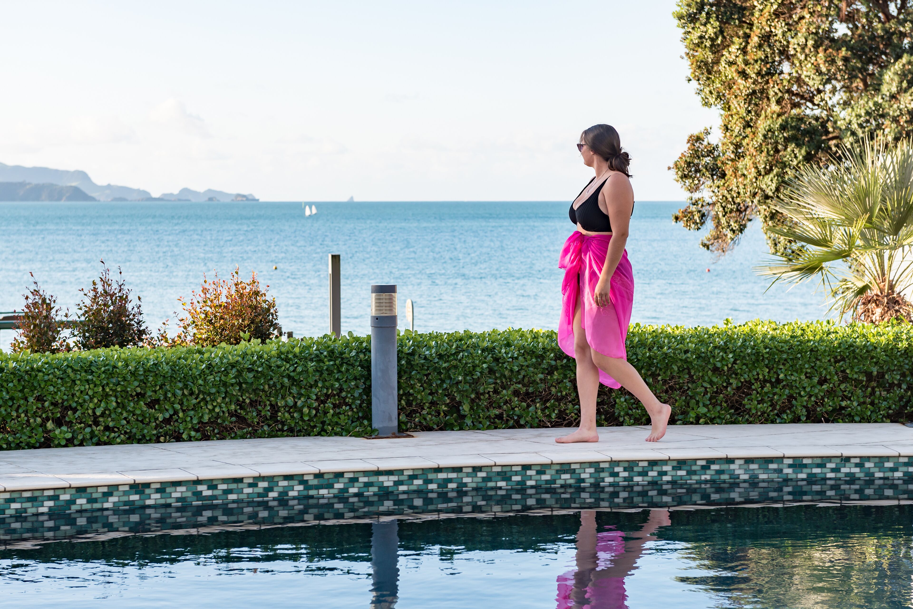 A poolside area with a view of the sea, a tiled edge, and a hedge.