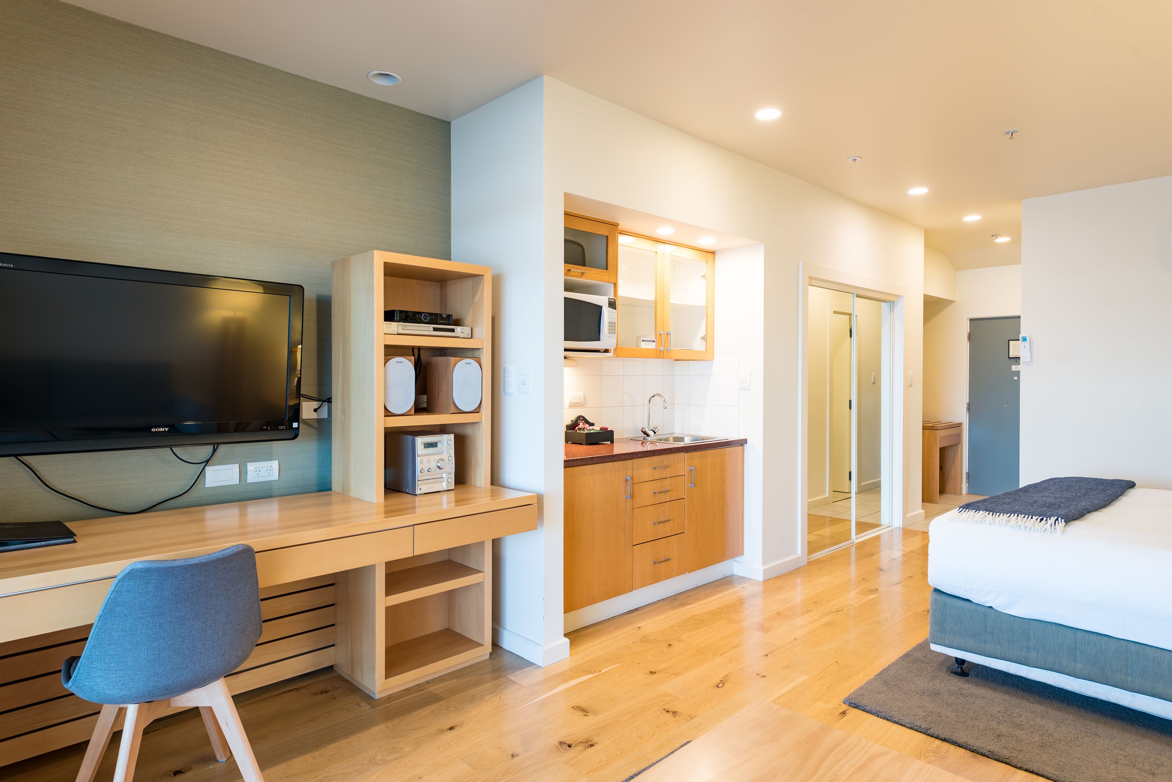 A modern hotel room with a wooden desk, a television, a kitchenette, and a bed.
