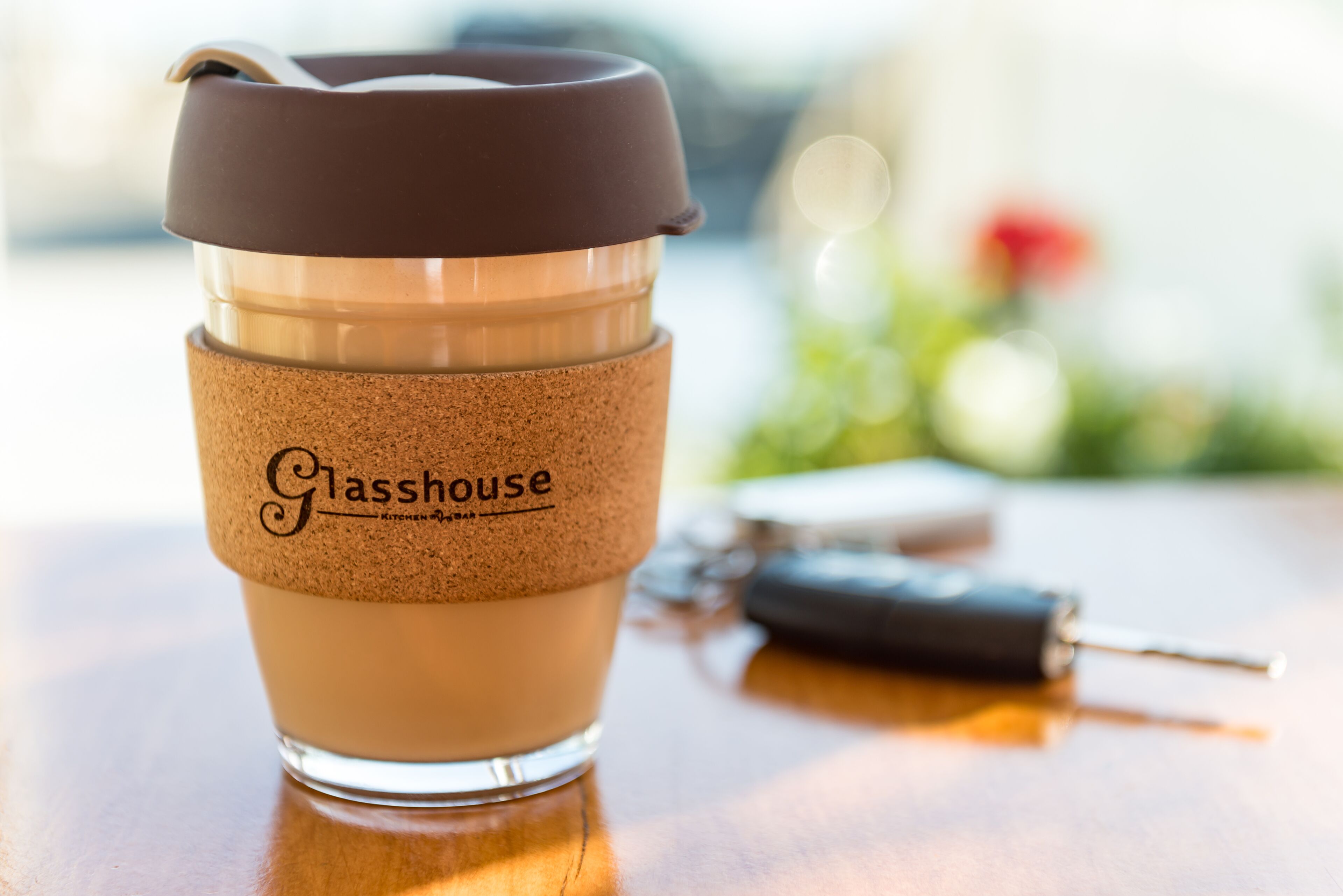 Eine Glaskaffeetasse mit Korkmanschette und braunem Deckel, beschriftet mit „Glasshouse“, auf einer Holztischfläche, in der Nähe ein Schlüsselanhänger.