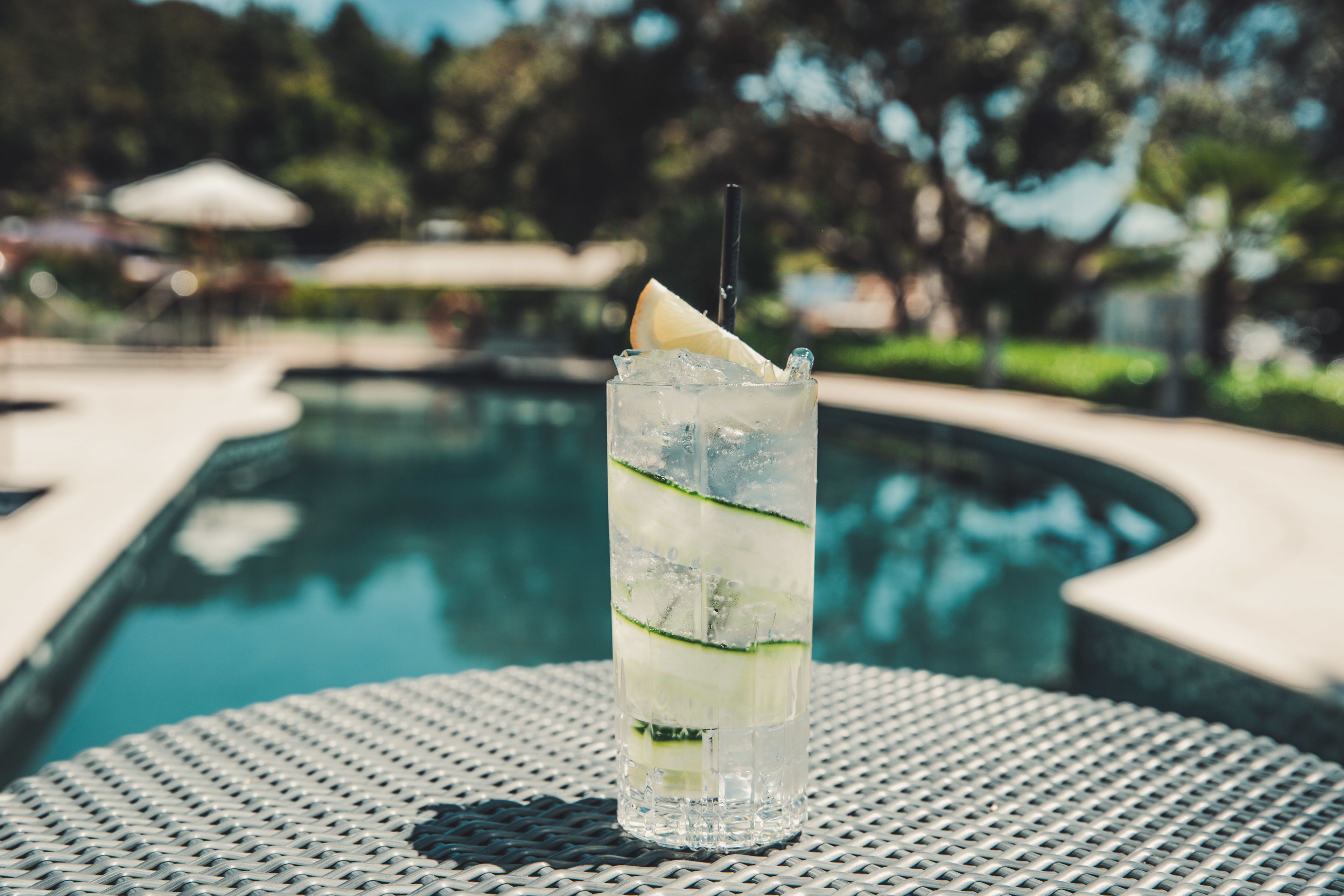 A glass of iced drink with a lemon slice and a straw, placed on a metal table near a pool.