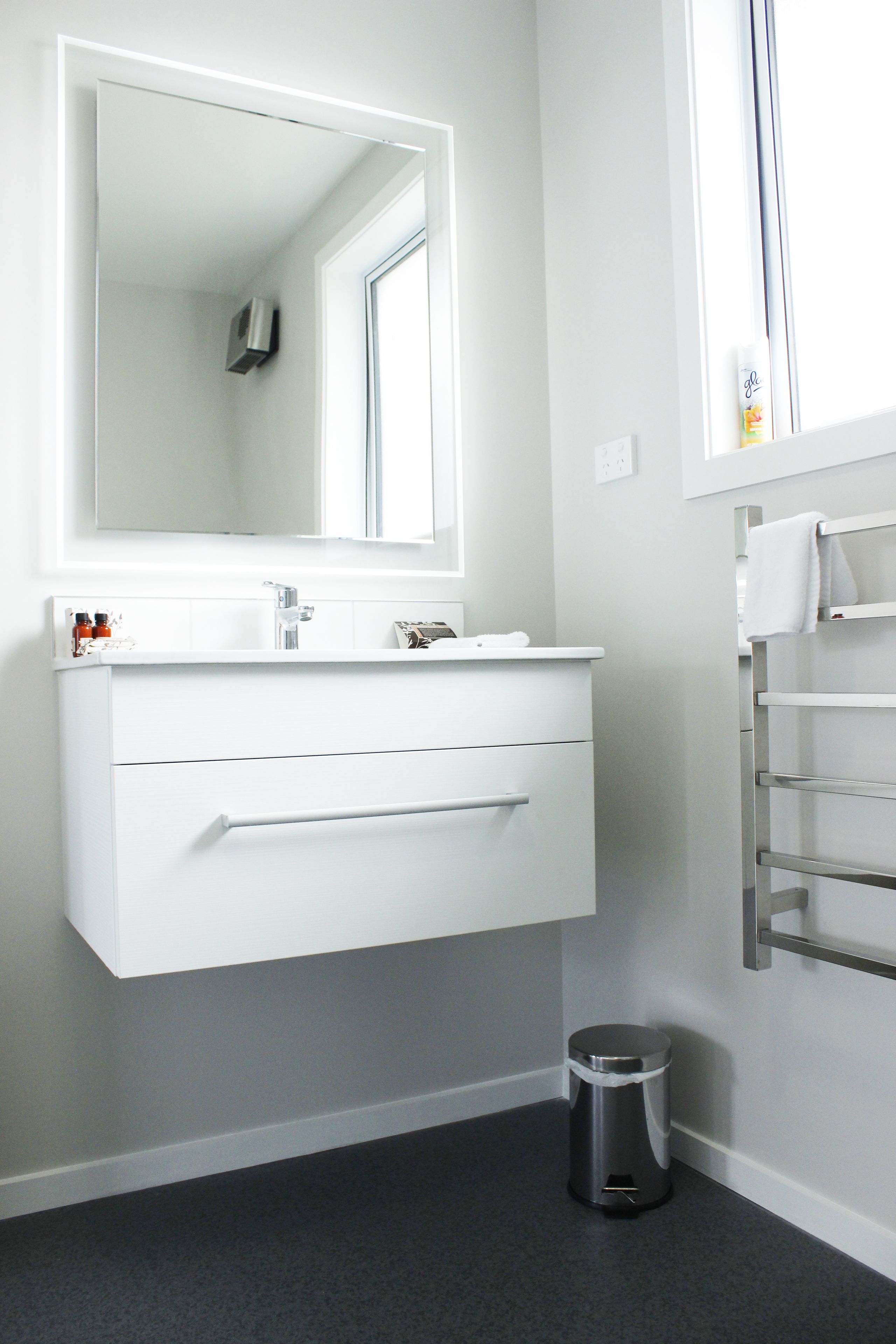 A modern bathroom with a white vanity, a large mirror, a towel rack, and a trash can.