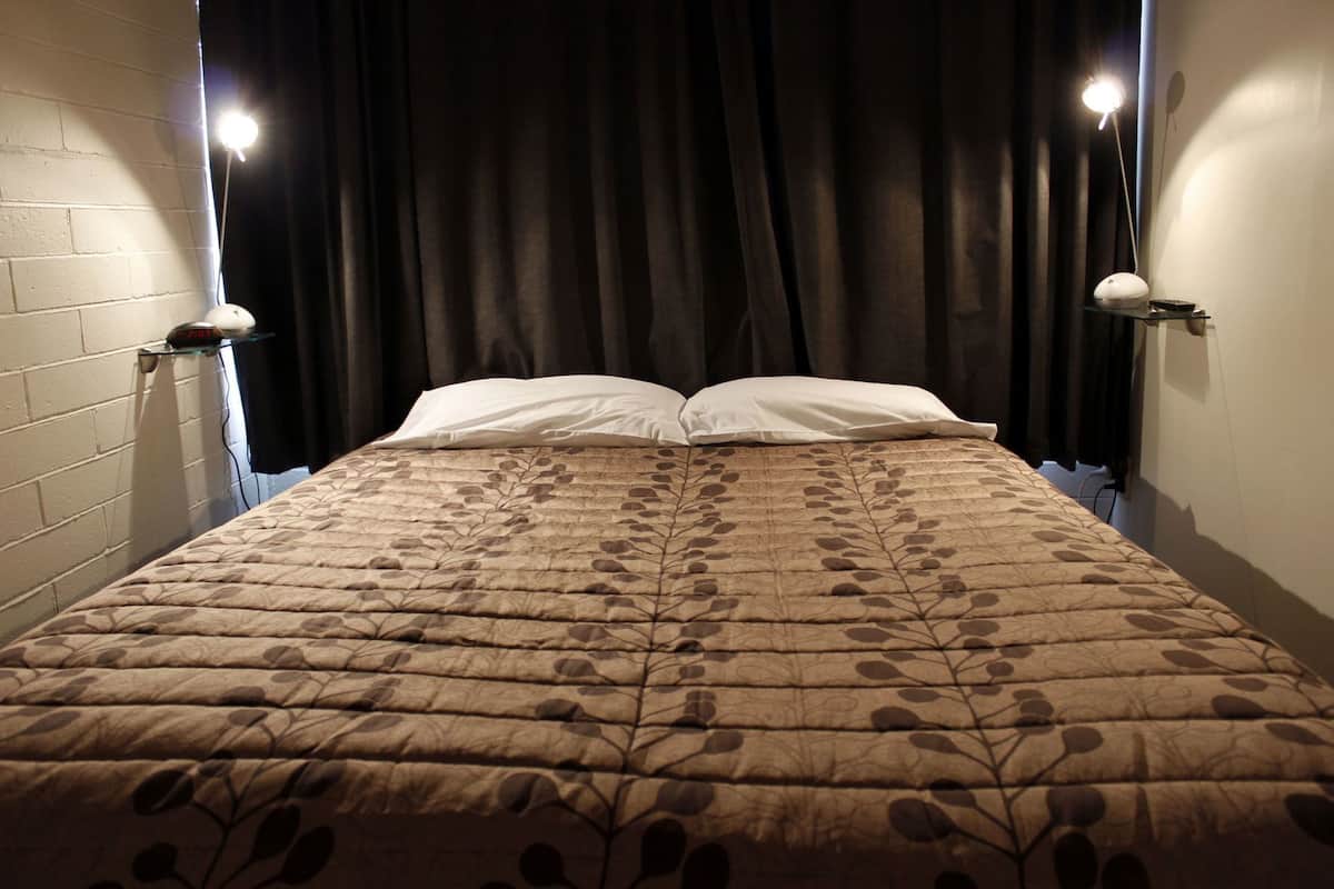 A bed with a leaf-patterned bedspread, two bedside lamps, and a dark curtain backdrop.