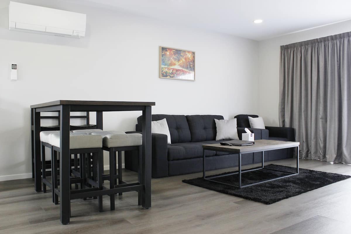 A modern living room with a dark sofa, a coffee table, and a small dining area with stools.