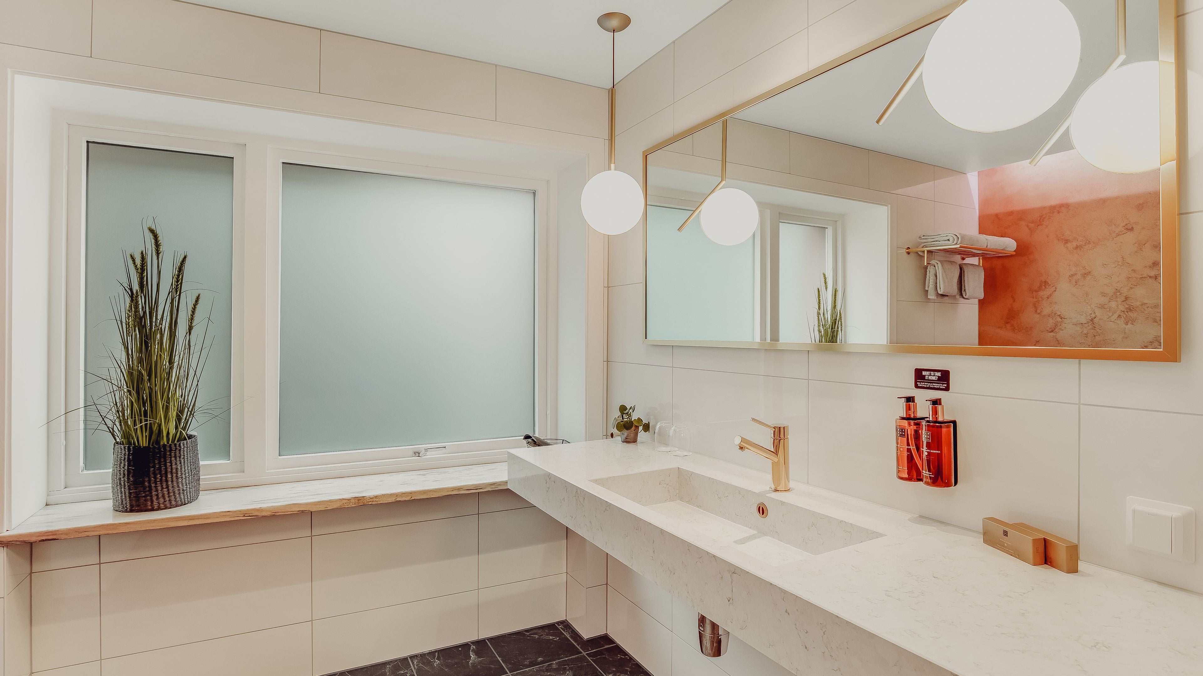 A modern bathroom with a large window, a white sink with a gold faucet, and a mirror with a gold frame.