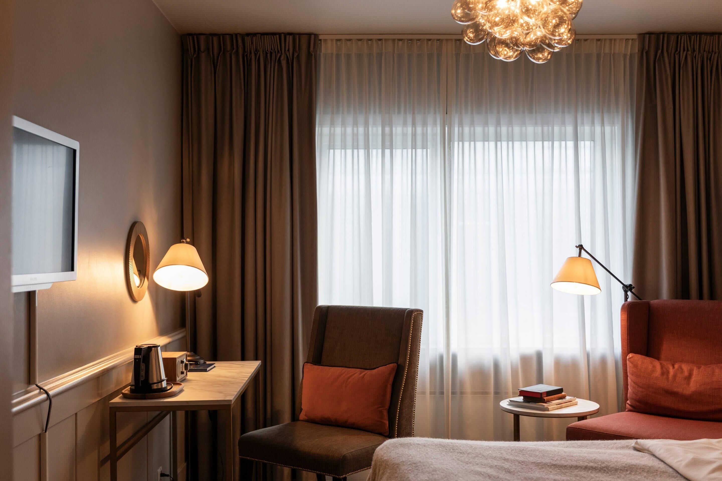 Habitación de hotel con cama, escritorio con lámpara, silla, televisor y ventana con cortinas.