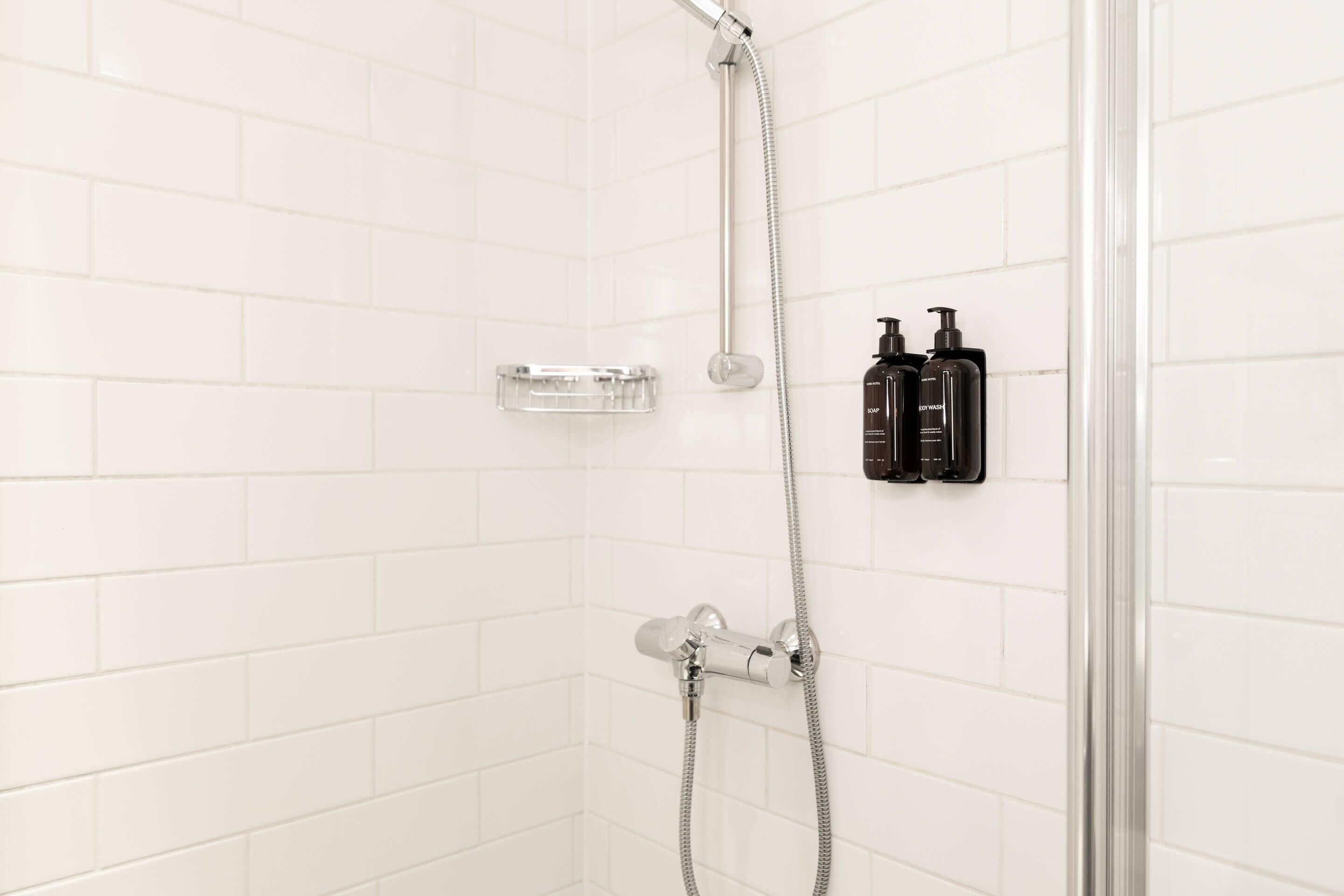 Un área de ducha con paredes de azulejos blancos, un plato de ducha de cristal y dos botellas negras en la pared.