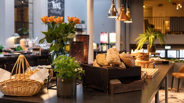 A well-lit indoor space with a long counter displaying bread, a vase with flowers, and a decorative lamp. - Home Hotel Grand Bodø (Bodo)