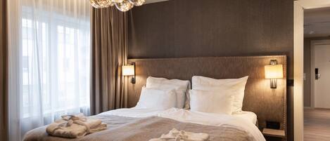 A hotel room with a large bed, two bedside lamps, a chandelier, and a view of the hallway.