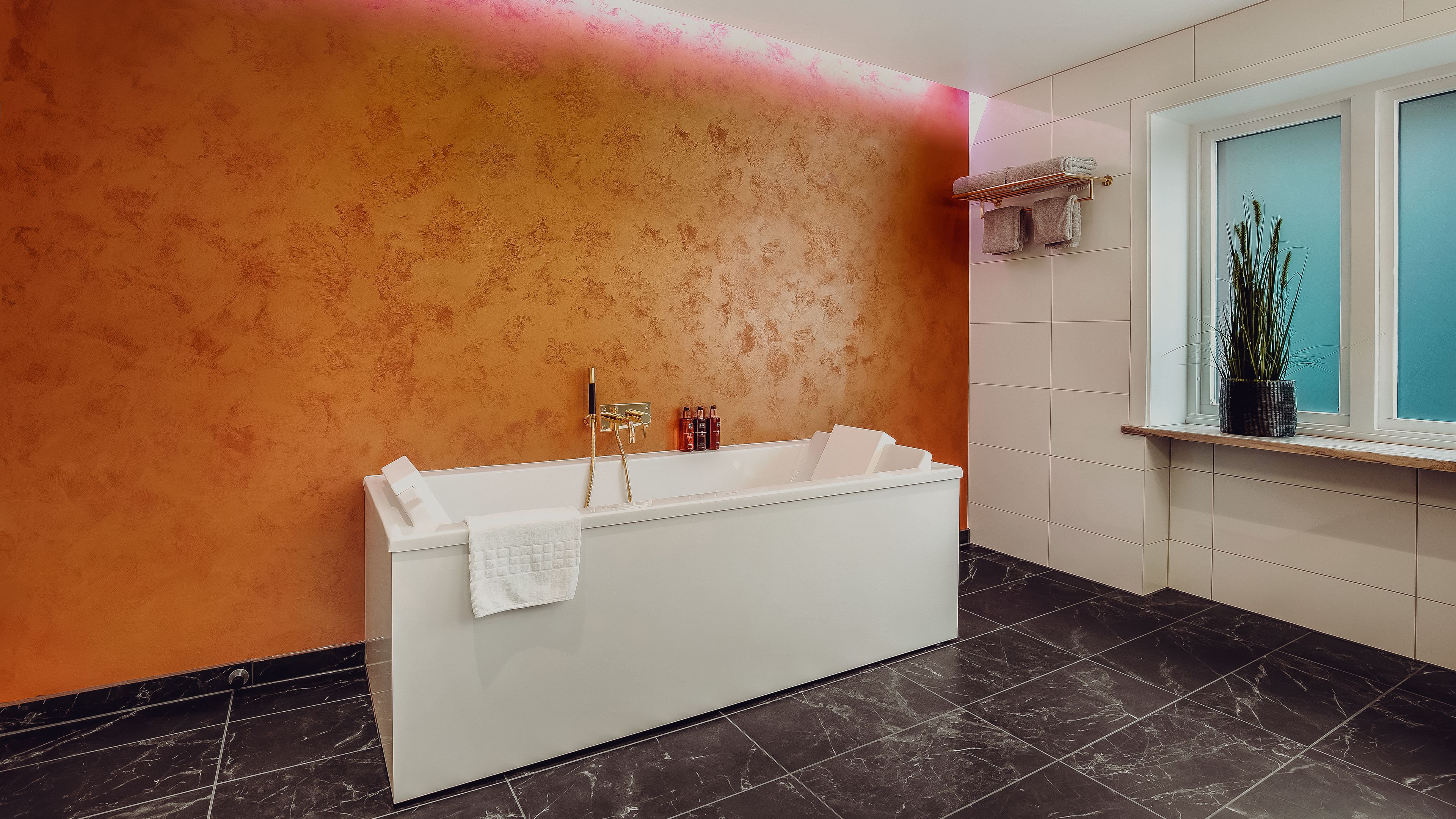 Un baño con bañera, perchero, ventana con planta y suelo de baldosas.