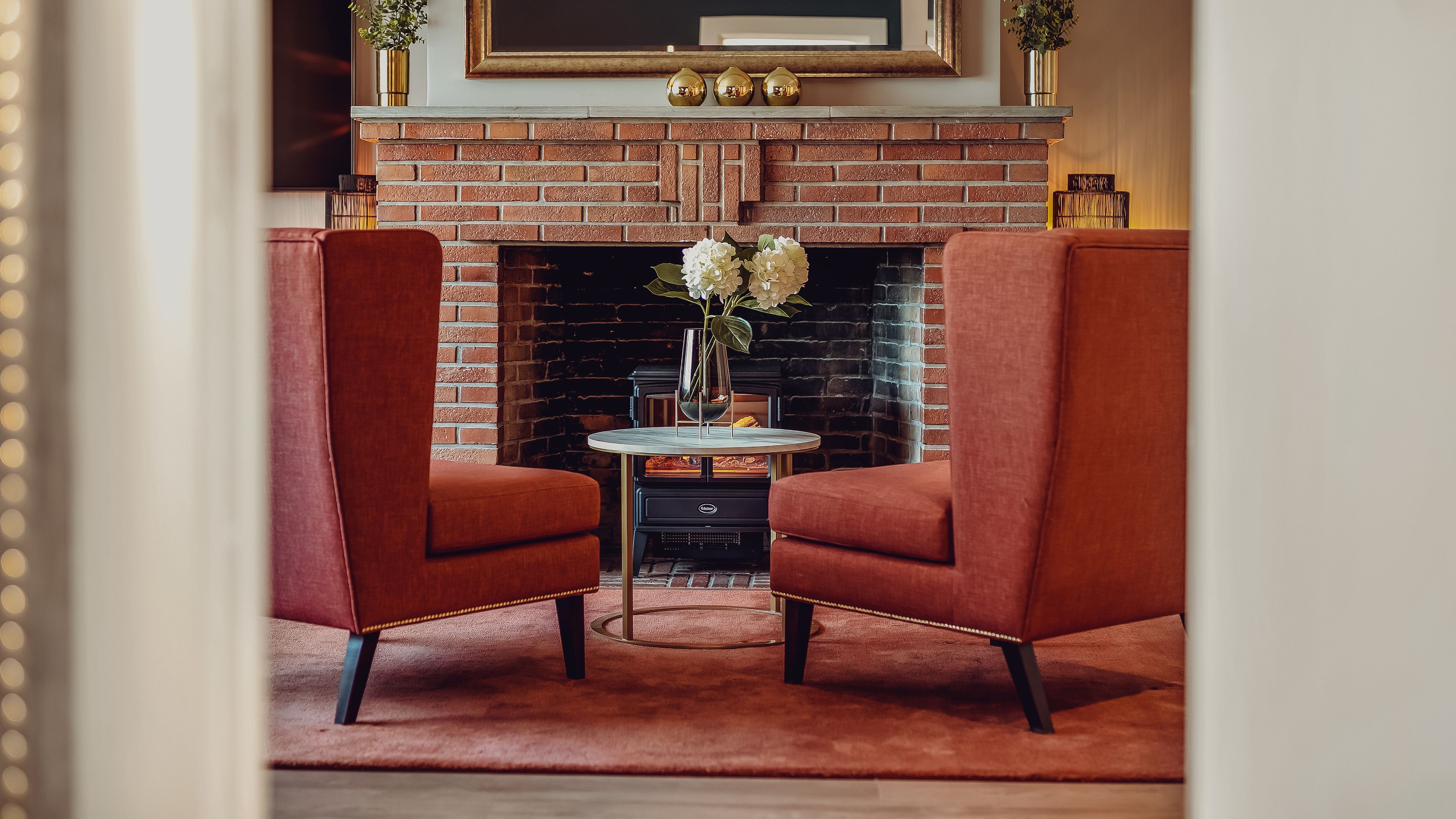 A cozy room with a brick fireplace, two red armchairs, a small round table with a vase of flowers, and a framed picture above the mantel.
