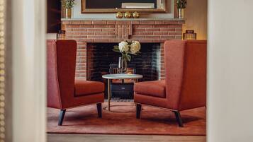 Una habitación acogedora con chimenea de ladrillo, dos sillones rojos, una mesita redonda con un jarrón de flores y un cuadro enmarcado sobre la repisa.