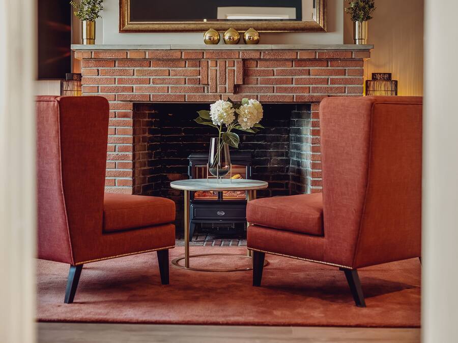 Una habitación acogedora con chimenea de ladrillo, dos sillones rojos, una mesita redonda con un jarrón de flores y un cuadro enmarcado sobre la repisa.