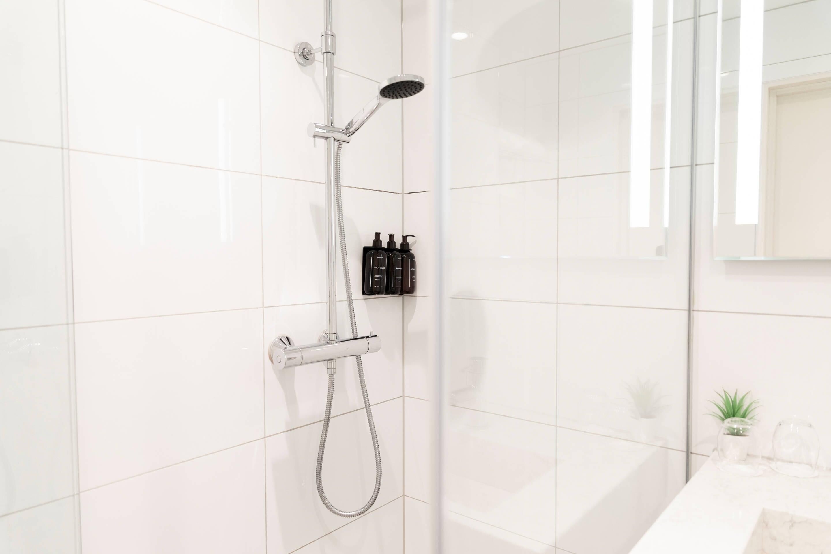 A modern bathroom with white tiles, a glass shower enclosure, a handheld showerhead, and a mounted handheld showerhead. There are two bottles of shampoo and conditioner on the wall.