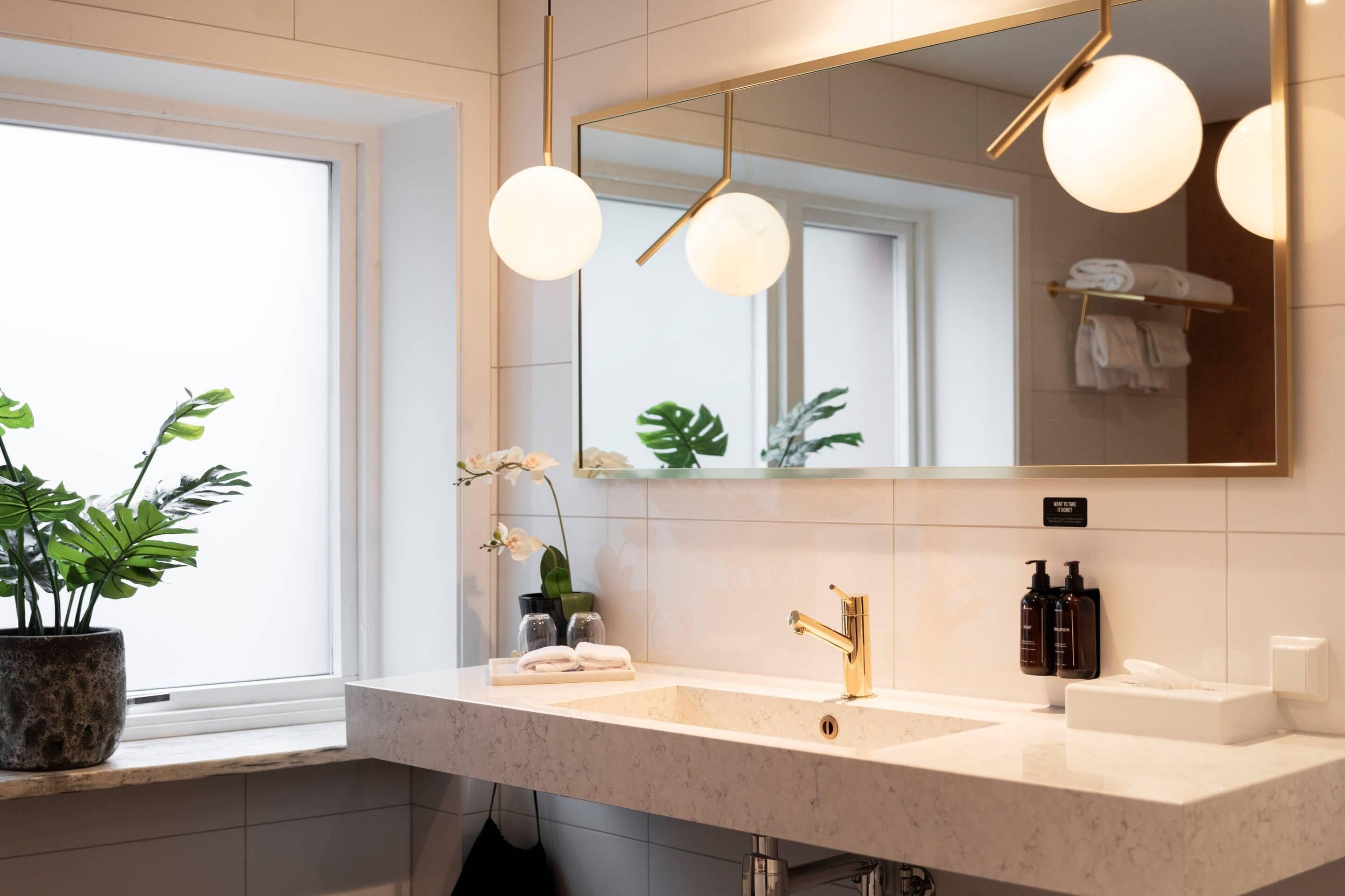 A modern bathroom with a large mirror, two sinks, a potted plant, and a window.