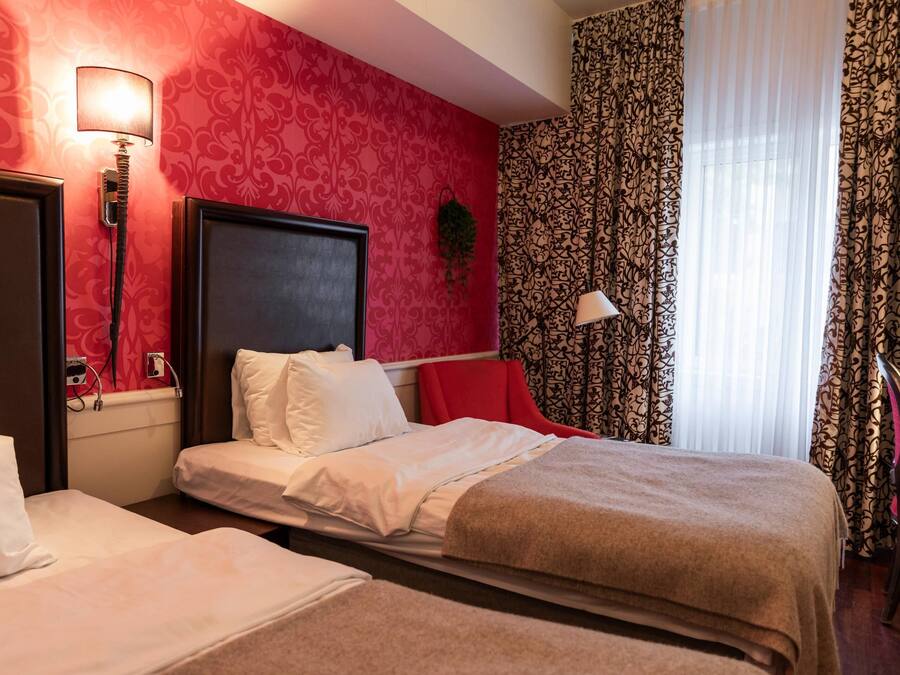 A hotel room with two beds, a red patterned wall, a window with patterned curtains, and a red chair.
