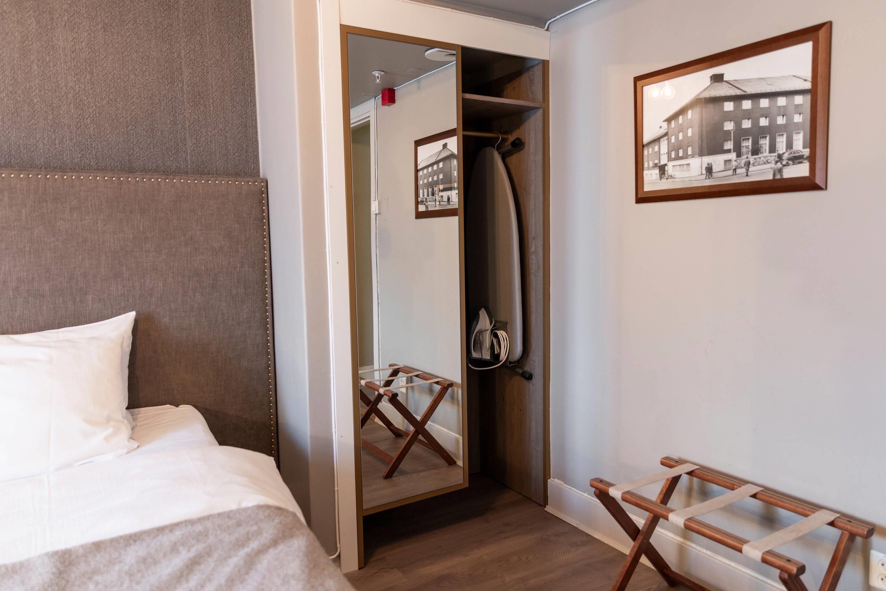 A hotel room with a bed, a wooden chair, a coat rack, a mirror, and a framed picture on the wall.