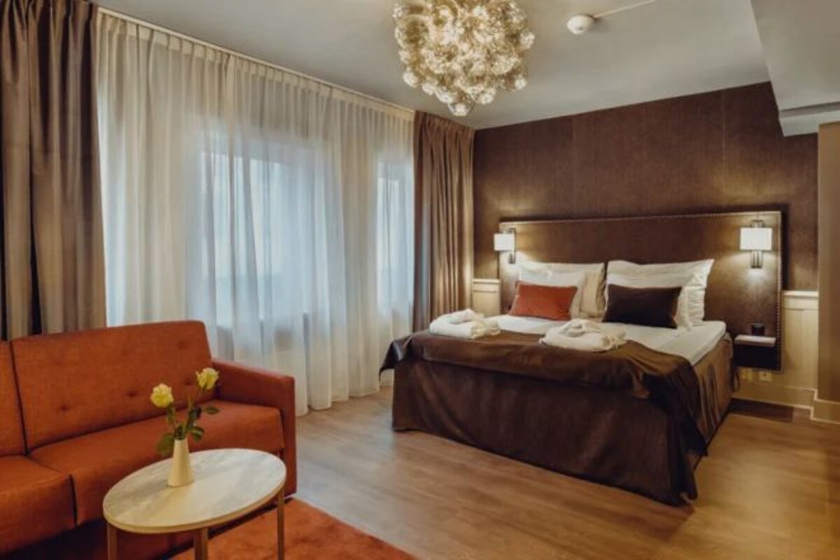 A hotel room with a large bed, a sofa, a round table with a vase, and a chandelier.