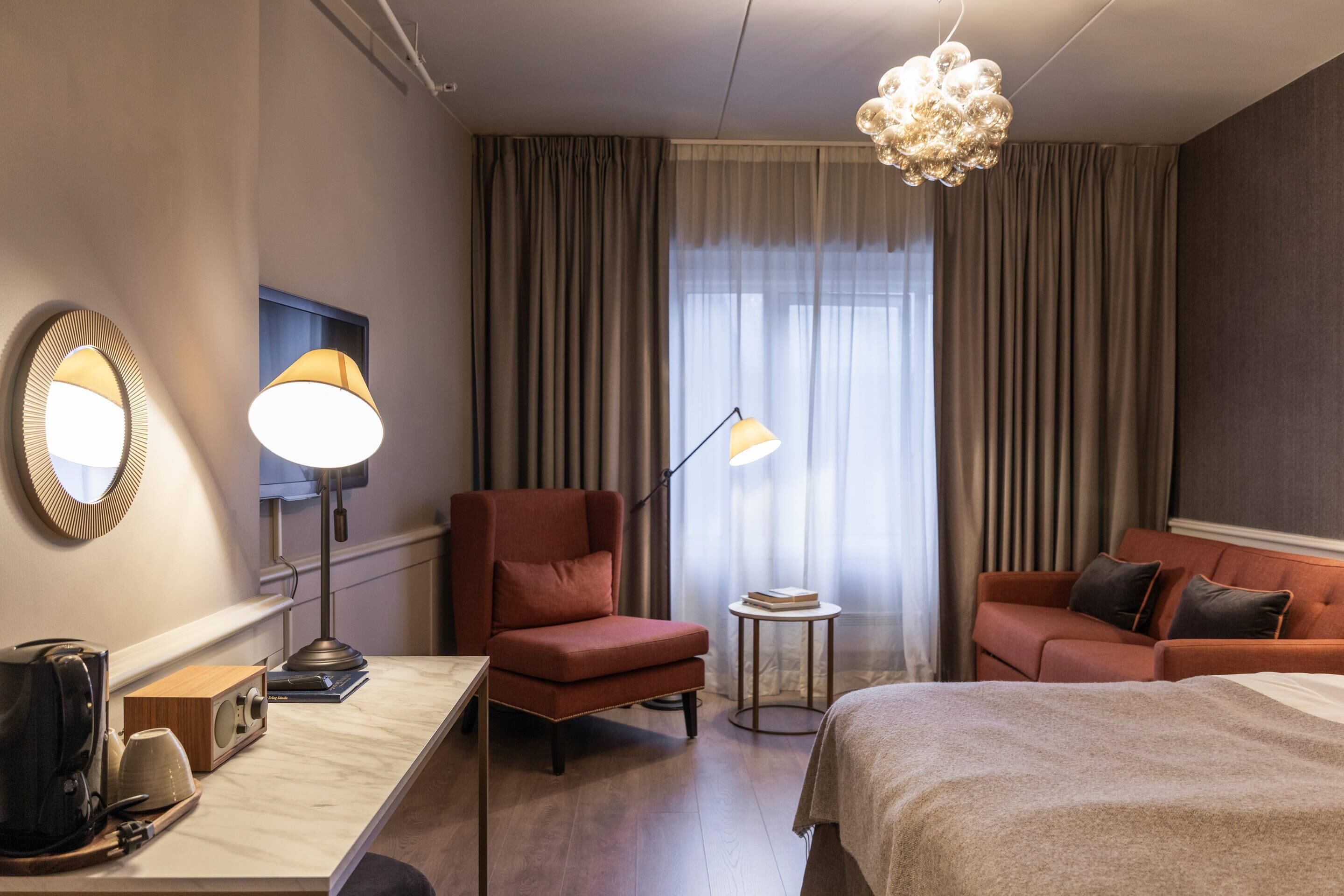 Habitación de hotel con cama, dos sillones, un escritorio con lámpara, espejo, una mesita con libros y un ventanal con cortinas.