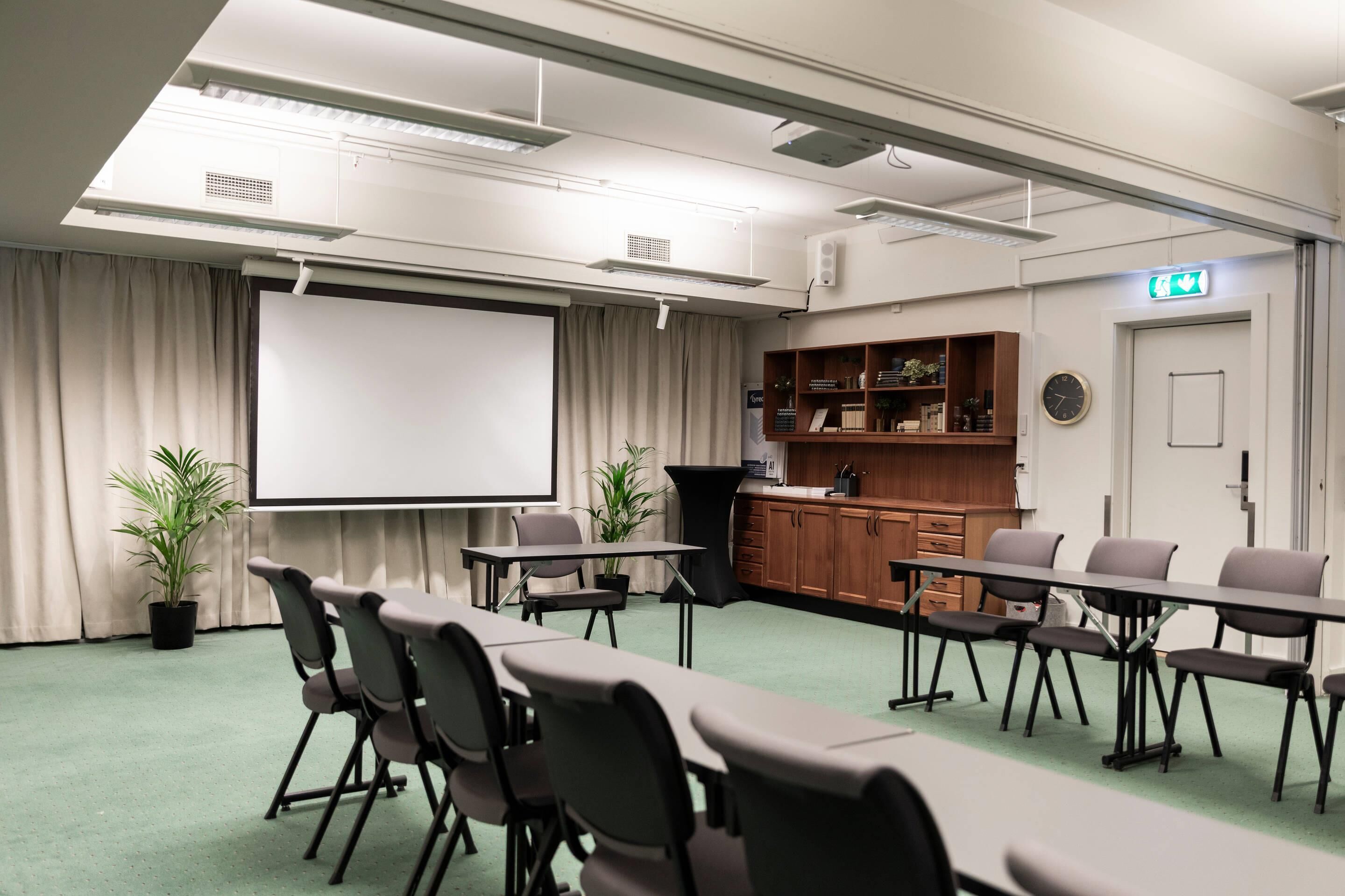 A conference room with a projector screen, tables, chairs, a kitchenette, and a clock on the wall.