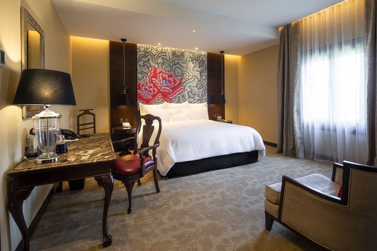 Ein Hotelzimmer mit einem Bett, einem Schreibtisch mit Lampe, einem Sessel und einem großen Wandteppich.
