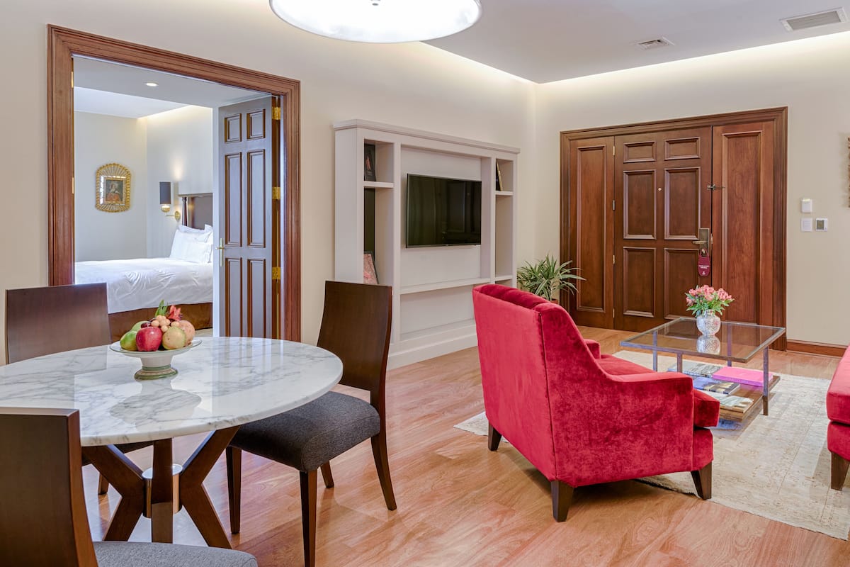 Ein Hotelzimmer mit einem Bett, einer Essecke mit einem Marmortisch, einem roten Sessel, einem an der Wand montierten Fernseher und Holztüren.